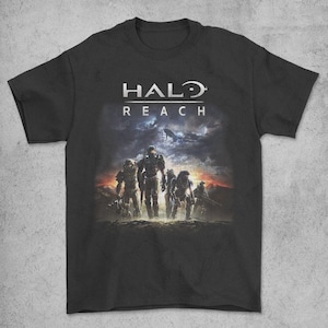 Peut inclure: T-shirt noir avec le logo Halo Reach en blanc. Le design comprend un graphisme de soldats en armure, sur fond de ciel dramatique avec un vaisseau spatial. Il s'agit d'une image promotionnelle du jeu vidéo.