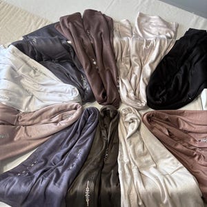 May include: A selection of hooded scarves in shades of black, brown, grey, and beige. Each scarf features a line of sparkling embellishments. The scarves are displayed in a circular arrangement, highlighting their diverse colours and textures.