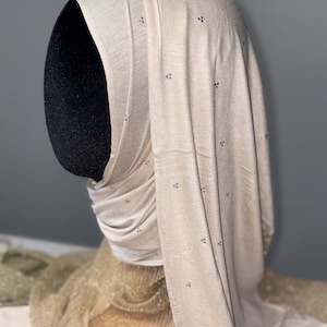 May include: A beige hijab, displayed on a black mannequin head. The fabric features small, dark decorative elements. The headscarf is arranged to highlight its form and texture, with gentle folds and a slight lustre.