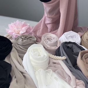 May include: An assortment of rolled and draped hijabs in a range of neutral tones, such as black, white, grey, and pinks and beiges. Some hijabs feature small, glittering details. A pink flower and rope are used as decorative elements.