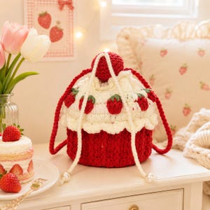 May include: A round, crocheted drawstring bag shaped like a cupcake with a deep red base and a white, ruffled top. The bag features small, red, crocheted strawberry appliqués around the white section and a red pom-pom on top with white cords.