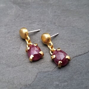 May include: A pair of gold earrings with a teardrop-shaped, faceted, dark red gemstone dangling from each earring.