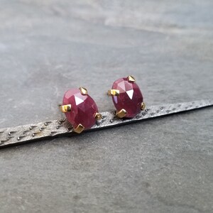 May include: A pair of ruby earrings with gold settings. The earrings are oval shaped and have a faceted cut. They are set on a dark gray textured surface.