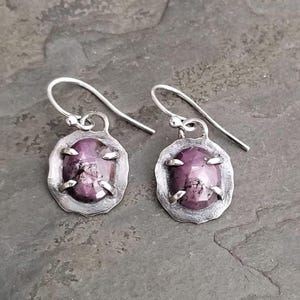 Könnte beinhalten: Silberne Ohrringe mit einem dunkelvioletten, ovalen Stein. Der Stein ist in einem strukturierten Silberrahmen mit vier Krappen gefasst. Die Ohrringe haben ein einfaches Haken-Design.