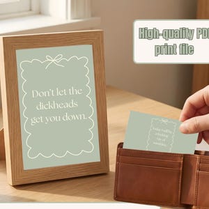 May include: A wooden framed print with the text "Don't let the dickheads get you down." A small card with the text "Today I will be a fucking ray of sunshine" is held near a brown leather wallet. The image also includes the text "High-quality PDF print file."