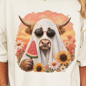 May include: An off-white t-shirt featuring a graphic of a highland cow wearing a white ghost sheet, round sunglasses, and holding a slice of watermelon. The cow is surrounded by sunflowers and daisies against a sunset-colored background.