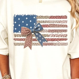 Op de afbeelding: Crèmekleurig T-shirt met een versleten Amerikaans vlagontwerp en een luipaardprint strik. De vlag heeft een blauw vierkant met witte sterren en rode en luipaardprint strepen. Het shirt heeft korte mouwen.