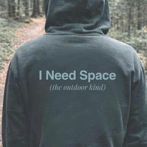 Könnte beinhalten: Ein dunkelgrüner Kapuzenpullover mit dem Text "I Need Space (the outdoor kind)" auf dem Rücken. Die Person steht auf einem Feldweg in einem Wald, umgeben von Bäumen. Der Kapuzenpullover hat eine Kapuze mit Kordelzug.
