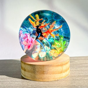 May include: A clear resin sphere with a diver figure and colorful coral reef design. The sphere sits on a round wooden base. The coral features shades of red, yellow, pink, and green, set against a blue background.