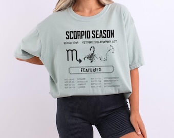 May include: A pale green t-shirt featuring the words "SCORPIO SEASON" and a scorpion illustration. The shirt lists dates and traits associated with the Scorpio zodiac sign. The shirt is worn with black shorts.