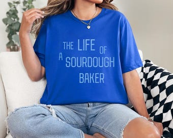 May include: Royal blue t-shirt with the text "THE LIFE OF A SOURDOUGH BAKER" in light blue. The shirt is worn with light blue jeans. The person is sitting on a white cushion with a black and white patterned pillow.