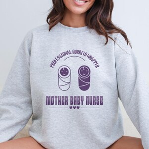 Puede incluir: Una sudadera gris claro con el texto "PROFESSIONAL BURRITO WRAPPER" arqueado sobre dos bebés envueltos en púrpura. Debajo, se muestra el texto "MOTHER BABY NURSE", con tres corazones debajo.