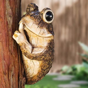 May include: A whimsical frog sculpture, in shades of brown and tan, is affixed to a tree trunk. The frog has a textured, detailed design, with one eye visible. The frog appears to be peeking out from behind the tree.