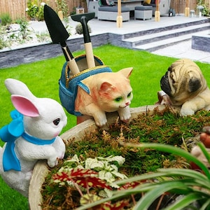 Könnte beinhalten: Ein Gartenpflanzer mit drei Tierfiguren: ein weißes Kaninchen mit blauer Schleife, eine orangefarbene Katze und eine Möpse. Die Katze hält eine blaue Tasche mit Gartengeräten. Der Pflanzer ist mit Grünzeug gefüllt.