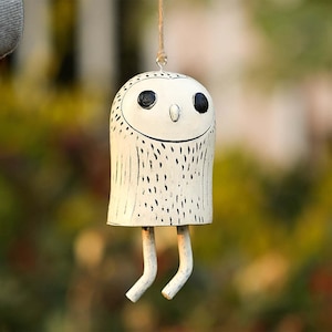May include: A handcrafted, white owl wind chime with black eyes and a smiling face. The owl has a cylindrical body with vertical etched lines and small, curved legs. It hangs from a natural twine string, creating a charming, rustic aesthetic.