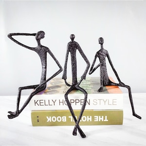 May include: Three black metal sculptures of stylized human figures in different poses. The figures are seated on books. The sculptures have a textured, dark finish against a white background. The books have text.