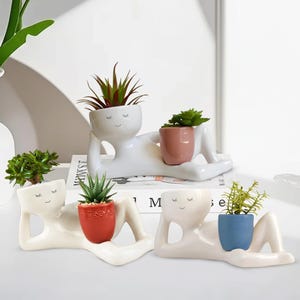 Peut inclure: Des pots de fleurs en céramique blanche en forme de personne allongée, chacun tenant une petite plante en pot. Les pots ont un design minimaliste avec des yeux fermés et une bouche souriante. Les pots sont de différentes couleurs, dont le rouge, le rose et le bleu.