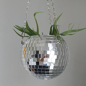 May include: A hanging disco ball planter with a chain. The spherical planter is covered in small, mirrored squares that reflect light. Green plants emerge from the top, adding a natural element to the decorative item.