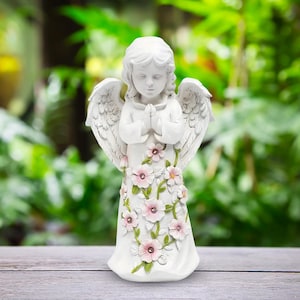 Può includere: Una statuina di angelo bianca con le mani giunte e gli occhi chiusi. L'angelo ha grandi ali dettagliate ed è adornato con accenti floreali rosa e bianchi. La statua è ambientata su uno sfondo verde sfocato.