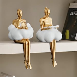 May include: Two gold-colored figurines of seated figures with white cloud-shaped bases. One holds a clear orb, the other a book. The figurines are perched on a white shelf, adding a touch of elegance to any space.