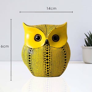 Peut inclure: Une figurine de hibou jaune, de 16 cm de haut et 14 cm de large, avec des motifs de points noirs. Le hibou a de grands yeux sombres et un bec. Une petite plante succulente en pot est en arrière-plan.