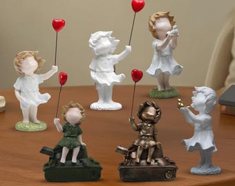 May include: A collection of decorative figurines depicting children. Some hold red heart-shaped balloons, while others hold birds. The figurines are in various colors, including white, bronze, and green, and are placed on a wooden surface.