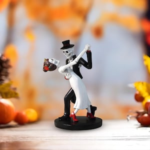 Pode incluir: Uma estatueta de Halloween que retrata um casal de esqueletos dançando. O esqueleto masculino usa um smoking preto e cartola, enquanto o esqueleto feminino está em um vestido branco e sapatos vermelhos. A base é preta, com um fundo desfocado com cores de outono.