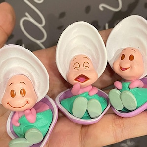 Peut inclure: Trois figurines de Tweedledee et Tweedledum d'Alice au pays des merveilles. Chaque figurine a un visage rose, un chapeau blanc et porte une robe verte. Elles sont assises dans une base en forme de coquillage rose et violet.