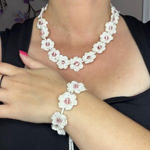 May include: A white crochet necklace and bracelet set. The jewelry features a floral design with pink accents. The necklace is around the neck, and the bracelet is on the wrist. The background is a black top.