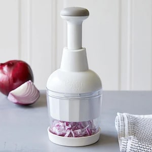 May include: A white onion chopper with a gray handle and a clear container filled with chopped red onion. A whole red onion and a slice of red onion are visible to the left. The text "Pampered Chef" is printed on the chopper.