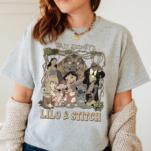 May include: A light gray t-shirt featuring a graphic of characters from the Disney movie Lilo & Stitch. The graphic includes the text "Wait Disney's Lilo & Stitch" in a brown font. The shirt is short-sleeved and has a crew neck.