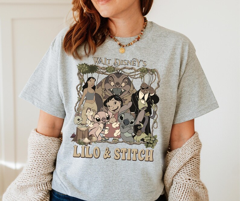 May include: A light gray t-shirt featuring a graphic of characters from the Disney movie Lilo & Stitch. The graphic includes the text "Wait Disney's Lilo & Stitch" in a brown font. The shirt is short-sleeved and has a crew neck.