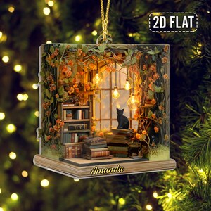 May include: A flat, square-shaped hanging ornament featuring a printed illustration of a cozy library nook with a black cat sitting on a stack of books, surrounded by orange flowers and hanging lights, with the name Amanda printed on the base.