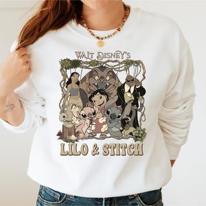 May include: White sweatshirt featuring a graphic of characters from Disney's Lilo & Stitch. The design includes the text "Walt Disney's Lilo & Stitch" in a retro font, with images of Lilo, Stitch, and other characters.