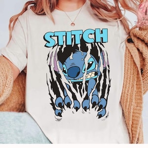 May include: A cream-colored t-shirt featuring the word "STITCH" in light blue block letters above a blue cartoon character. The character is breaking through a black, torn-paper effect, with its claws visible.
