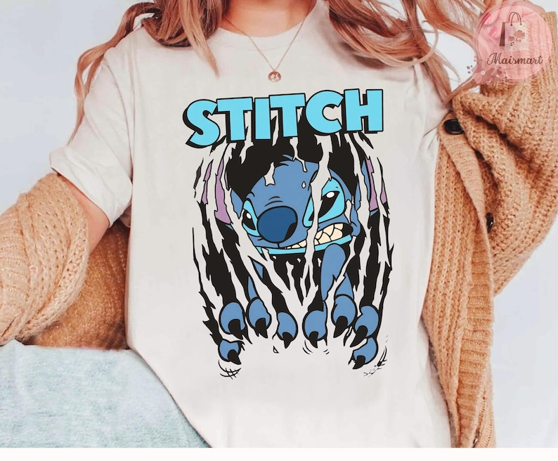 May include: A cream-colored t-shirt featuring the word "STITCH" in light blue block letters above a blue cartoon character. The character is breaking through a black, torn-paper effect, with its claws visible.