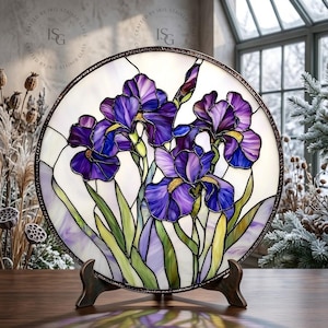 May include: A round stained glass panel featuring purple iris flowers with green leaves against a white background. The artwork is displayed on a bronze-colored stand. The panel has a dark brown border and is set in front of a window.