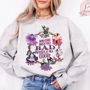 May include: A light gray sweatshirt featuring a colorful graphic of Disney villains. The graphic includes the text "HOW COME BEING BAD FEELS SO GOOD?" in a stylized font, surrounded by images of various villains.