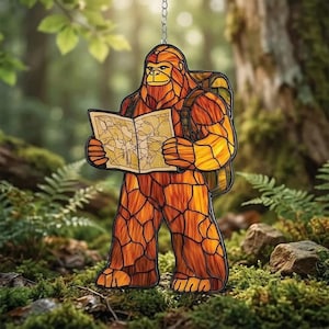 May include: A stained glass Bigfoot figure with a backpack, holding a map. The figure is primarily orange and brown, with yellow highlights. It is hanging from a chain, set against a blurred forest background.