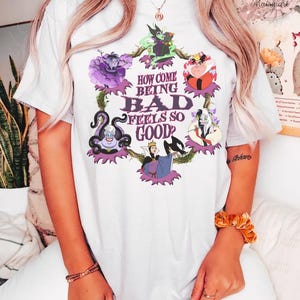 May include: White t-shirt featuring a colorful graphic of Disney villains, including Ursula, the Evil Queen, and Cruella de Vil. The text "HOW COME BEING BAD FEELS SO GOOD?" is printed in the center of the design. The shirt is made of a soft material.