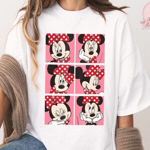 Puede incluir: Camiseta blanca con un dise&ntilde;o de seis paneles de Minnie Mouse en varias poses. Cada panel muestra a Minnie con su lazo rojo de lunares blancos. El fondo es rosa.