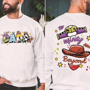 Puede incluir: Sudadera blanca con la palabra "DADA" en letras de bloque coloridas en la parte delantera, decorada con personajes de dibujos animados. La parte trasera presenta una nave espacial, un sombrero de vaquero y la frase "To infinity and beyond."