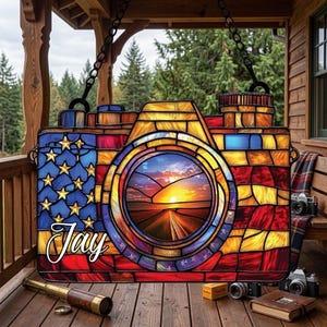 May include: A stained glass camera-shaped decoration featuring the American flag design. The lens displays a sunset scene. The name "Jay" is written in cursive. The decoration hangs from a chain and is displayed on a wooden porch.