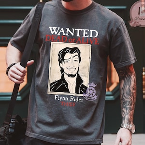 Puede incluir: Camiseta gris oscuro con un diseño de cartel de búsqueda. El cartel muestra una ilustración de dibujos animados de un hombre, con el texto "WANTED Dead or Alive" arriba y "Flynn Rider Thief" abajo. La camiseta tiene cuello redondo y mangas cortas.