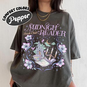 May include: A charcoal gray t-shirt featuring the text Midnight Book Club Reader in a stylized font. The graphic includes a gothic castle, a teacup, an open book, a candelabra, a crescent moon, and floral accents in shades of pink, blue, and white.