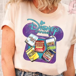 Peut inclure: T-shirt crème avec un motif Disney Channel. Le motif comprend le logo Disney Channel, des oreilles de Mickey et des images de films comme "High School Musical" et "Camp Rock". Le t-shirt a des manches courtes.