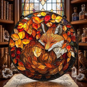 May include: Round stained glass window art featuring a sleeping fox nestled among autumn leaves. The fox has orange and white fur, surrounded by red, orange, and yellow leaves. The artwork is framed in black and hangs from a chain.