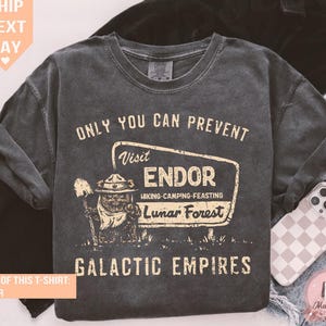 Puede incluir: Camiseta de manga larga color pimienta con un gráfico de estilo vintage. El gráfico dice "ONLY YOU CAN PREVENT Visit ENDOR Hiking-Camping-Feasting Lunar Forest GALACTIC EMPIRES". El diseño presenta un personaje de dibujos animados con una pala.