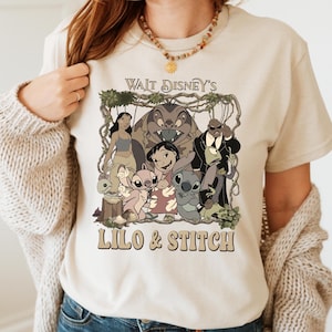 May include: Beige t-shirt featuring a graphic of characters from Disney's Lilo & Stitch. The design includes the text "Walt Disney's Lilo & Stitch" in a brown font. The image shows Lilo, Stitch, and other characters from the animated film.