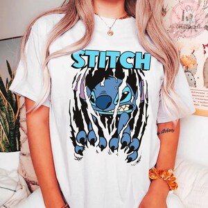 May include: White t-shirt featuring a blue cartoon character, Stitch, bursting through a torn design. The word "STITCH" is printed in blue above the character. The design has black and purple accents.